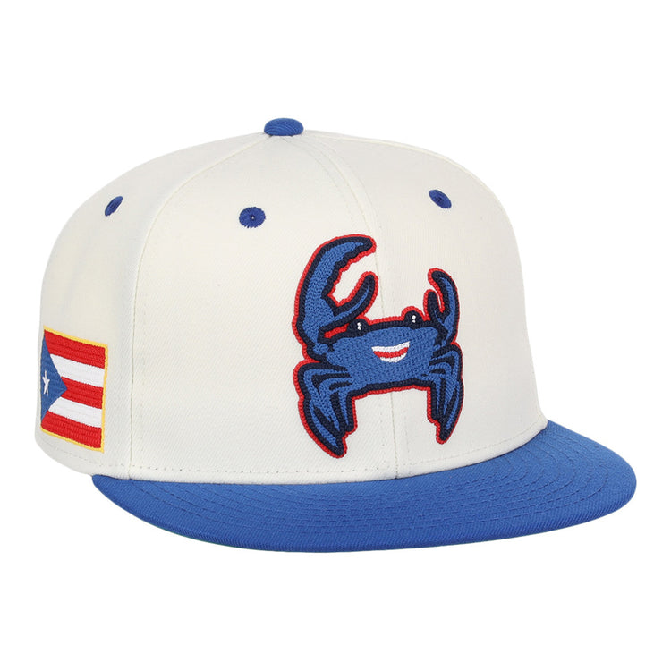 The Ebbets Field Santurce Cangrejeros Chainstitch Fitted Ballcap is a white cap with a blue brim, featuring a smiling blue crab patch on the front and a Puerto Rican flag patch on the side.