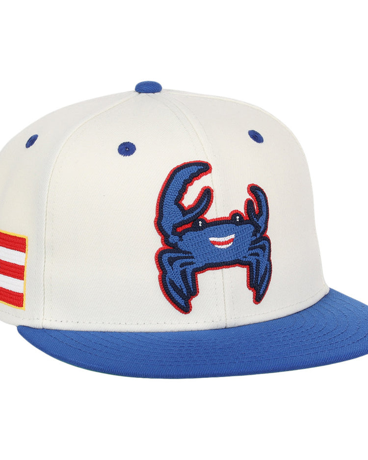 The Ebbets Field Santurce Cangrejeros Chainstitch Fitted Ballcap is a white cap with a blue brim, featuring a smiling blue crab patch on the front and a Puerto Rican flag patch on the side.
