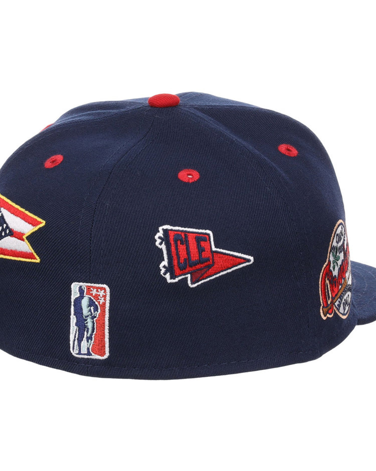 The Ebbets Field Cleveland Buckeyes NLB Varsity Fitted Ballcap is a navy blue cap featuring embroidered patches of the Ohio flag, a red CLE pennant, a baseball player silhouette, and a team logo.