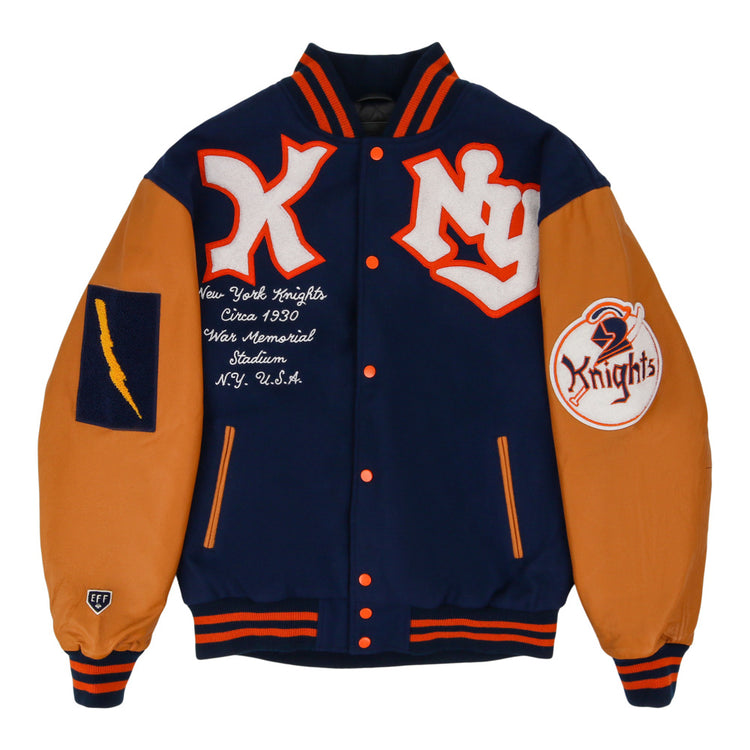 The Ebbets Field New York Knights Dna Varsity Jacket features navy blue and orange colors, NY and Knights patches, a lightning bolt patch on one sleeve, orange-trimmed collar, cuffs and hem, plus embroidered chest text referencing the New York Knights.