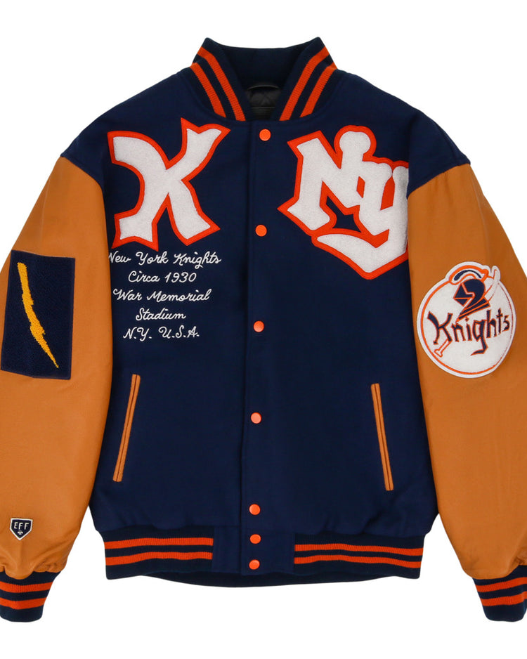 The Ebbets Field New York Knights Dna Varsity Jacket features navy blue and orange colors, NY and Knights patches, a lightning bolt patch on one sleeve, orange-trimmed collar, cuffs and hem, plus embroidered chest text referencing the New York Knights.