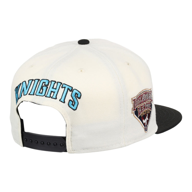 The Ebbets Field Charlotte Knights MiLB Snapback Ballcap features a white design with KNIGHTS embroidered in blue on the back, a HERITAGE HERALD side patch, a black brim, and an adjustable black strap.