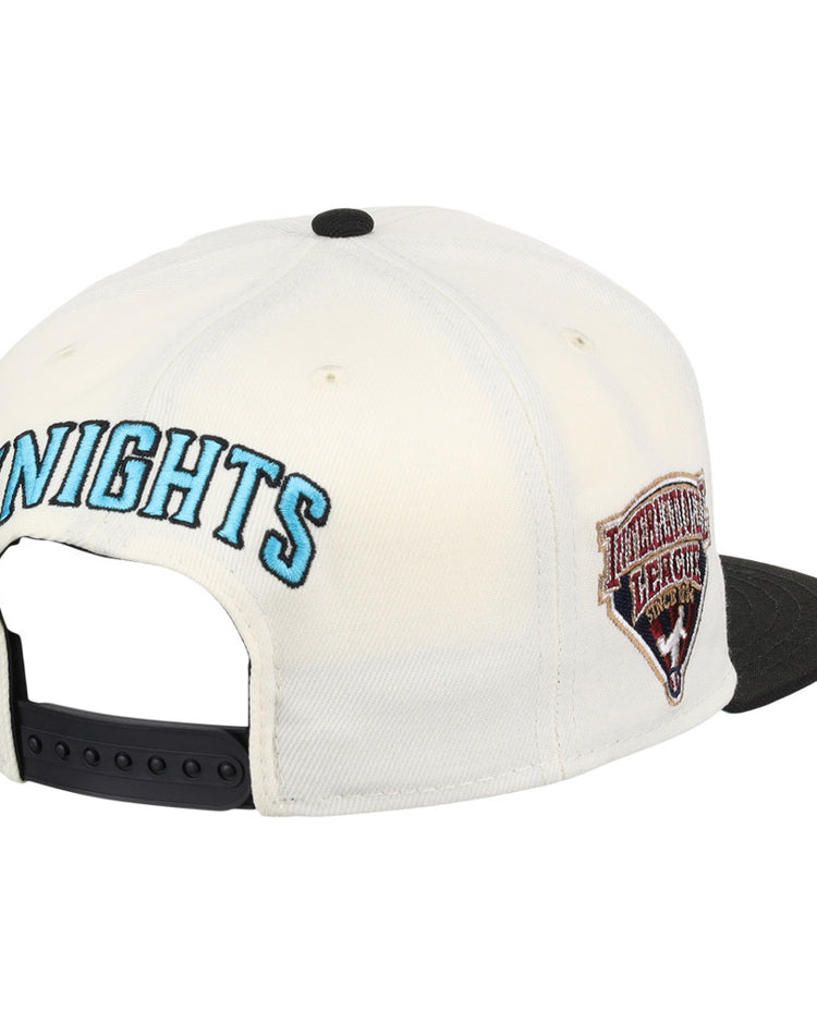 The Ebbets Field Charlotte Knights MiLB Snapback Ballcap features a white design with KNIGHTS embroidered in blue on the back, a HERITAGE HERALD side patch, a black brim, and an adjustable black strap.