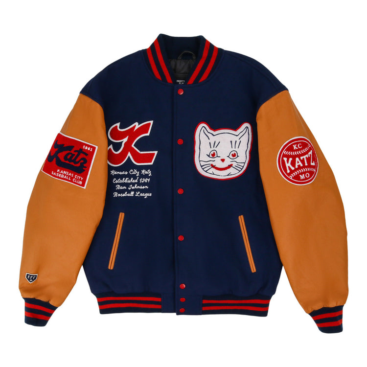 The Kansas City Katz Dna Varsity Jacket by Ebbets Field features navy blue body, tan sleeves, red and white striped cuffs, and embroidered patches including a smiling cat face, logos, and text on the chest and sleeves.