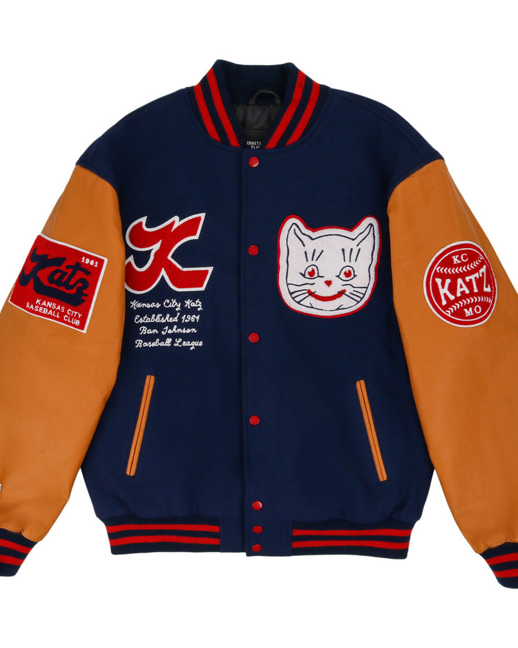 The Kansas City Katz Dna Varsity Jacket by Ebbets Field features navy blue body, tan sleeves, red and white striped cuffs, and embroidered patches including a smiling cat face, logos, and text on the chest and sleeves.