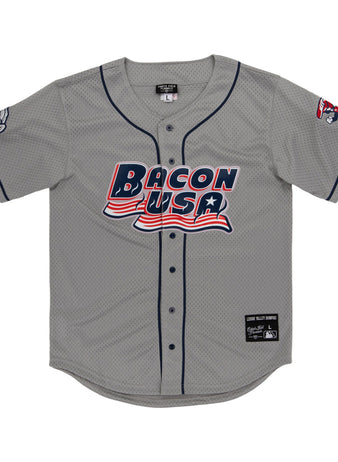 The Lehigh Valley Ironpigs MiLB Button Down Jersey by Ebbets Field is a gray baseball jersey with navy trim, “Bacon USA” in red, white, and blue on the front, plus cartoon bacon designs on the left sleeve and right shoulder.