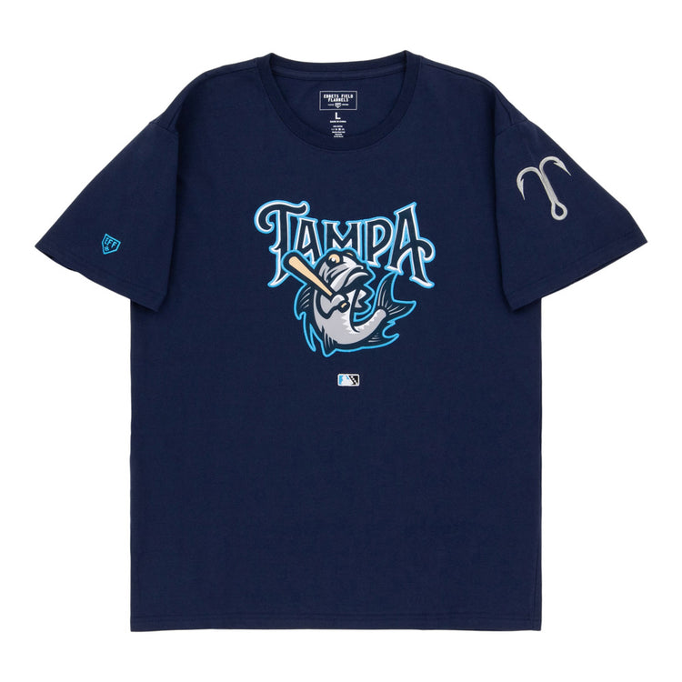 The Ebbets Field Tampa Tarpoons MiLB Fashion Cotton Short Sleeve Tee is a navy blue shirt with a stylized fish swinging a bat under the word “Tampa,” an MLB logo below, and a gray fishing hook on the left sleeve.