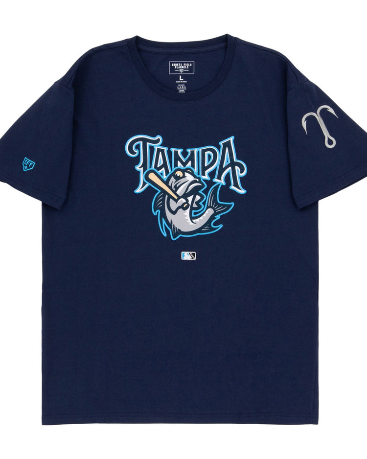The Ebbets Field Tampa Tarpoons MiLB Fashion Cotton Short Sleeve Tee is a navy blue shirt with a stylized fish swinging a bat under the word “Tampa,” an MLB logo below, and a gray fishing hook on the left sleeve.