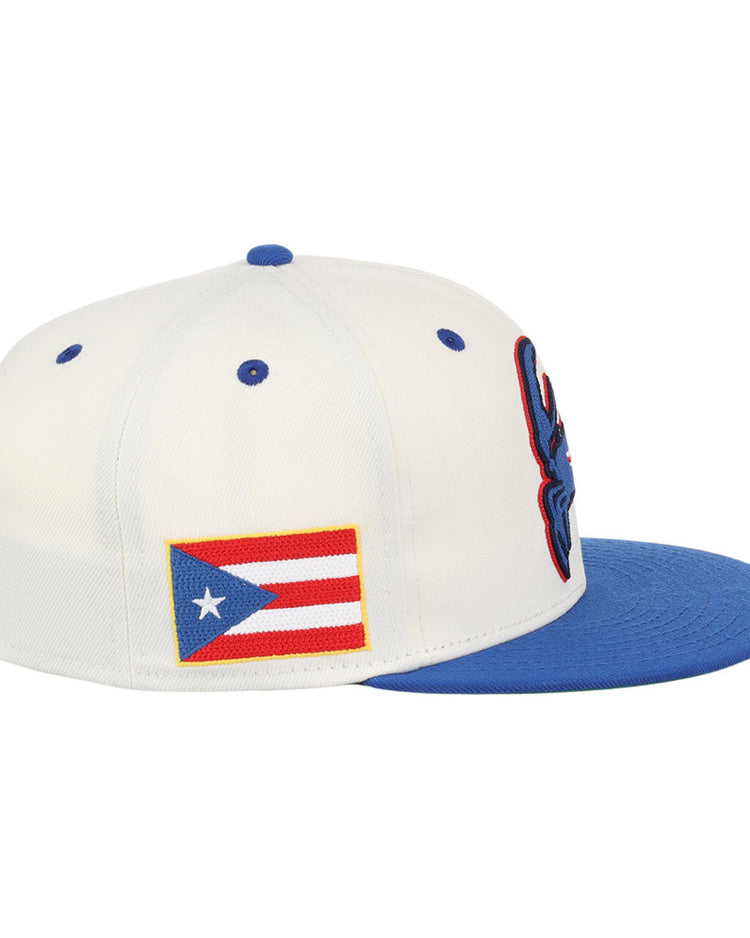 The Ebbets Field Santurce Cangrejeros Chainstitch Fitted Ballcap is a white hat with a blue brim, blue embroidered front details, and a Puerto Rican flag patch on the side.