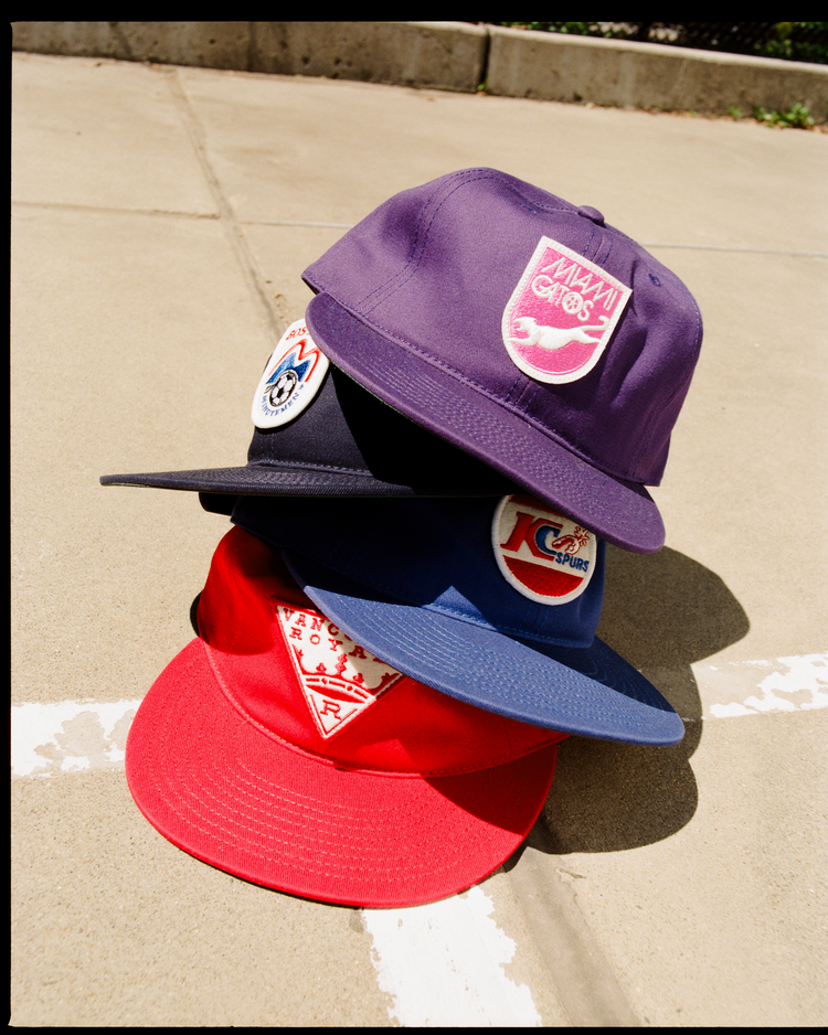 Four colorful Ebbets Field Miami Gatos Cotton Twill Ballcaps—purple, black, blue, and red—rest on sunlit concrete; each features unique embroidered patches and logos inspired by Miami Gatos and NASL heritage.