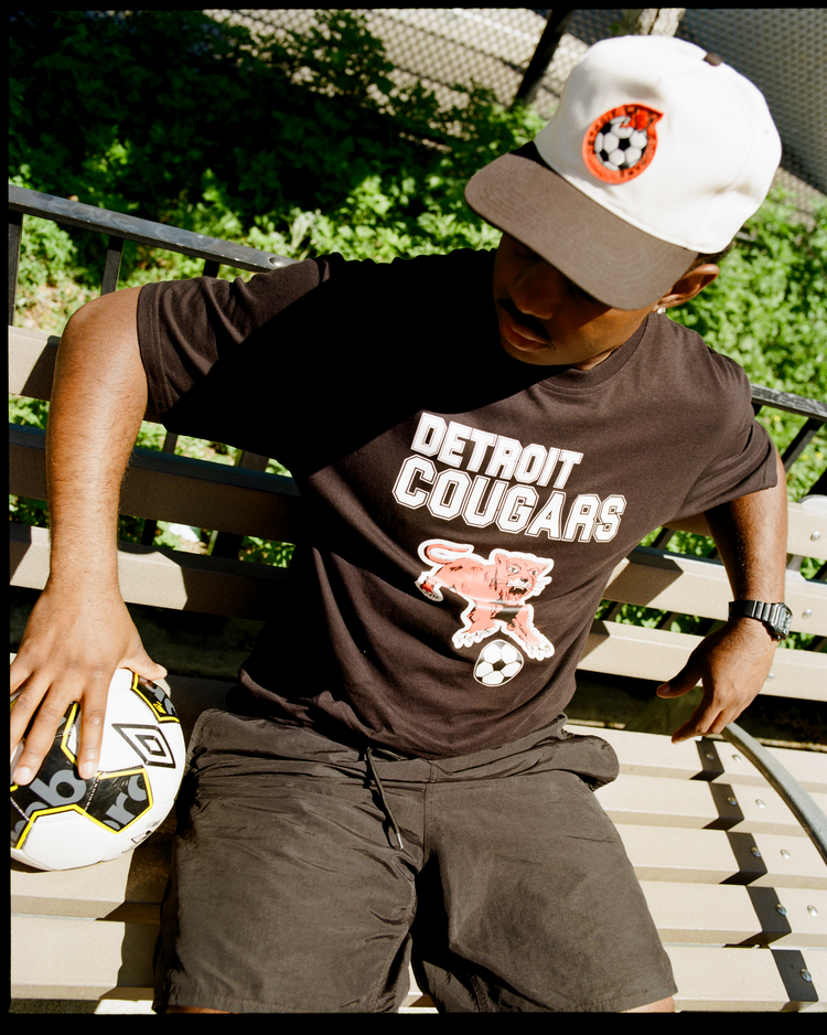 Wearing an Ebbets Field Detroit Cougars Short Sleeve Graphic T-Shirt and a soccer-themed cap, a person sits on a bench holding a soccer ball. Greenery and a fence can be seen in the background, evoking classic soccer vibes.