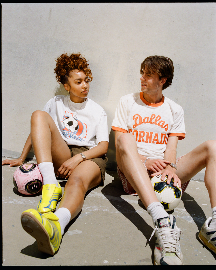 Two people sit against a light wall with soccer balls. The person on the right wears an Ebbets Field Dallas Tornado Mesh Soccer Jersey, while the other is in a white graphic tee, shorts, and neon sneakers.