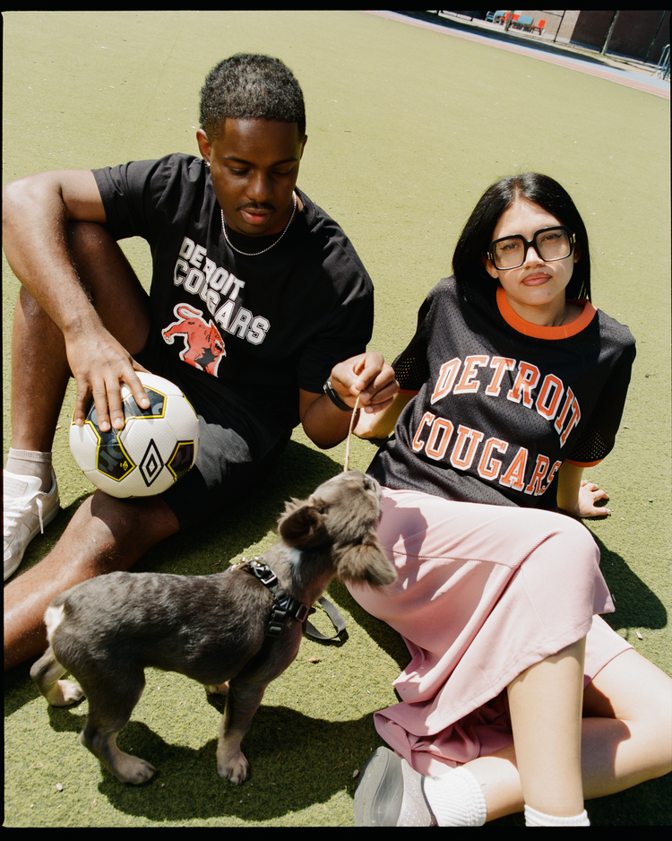 A man with a soccer ball and a woman in glasses and skirt play with their dog on the grass, both wearing Ebbets Field Detroit Cougars Mesh Soccer Jerseys.