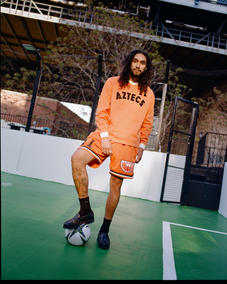 A man with long curly hair stands on a green court, resting one foot on a soccer ball. He wears an orange Ebbets Field Los Angeles Aztecs Vintage Soccer Goalie Jersey and shorts. A fence and trees are in the background.