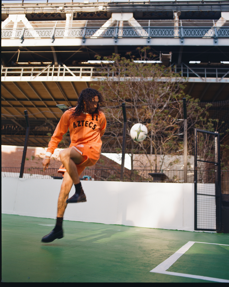 A person with long hair in an Ebbets Field Los Angeles Aztecs Vintage Soccer Goalie Jersey and black shoes kicks a soccer ball mid-air on an outdoor court, with a metal bridge and trees in the background.