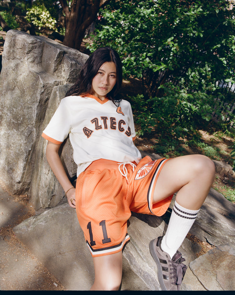 A person with long dark hair sits on a rock outdoors, wearing Ebbets Field Los Angeles Aztecs Mesh Soccer Shorts, a white jersey, striped socks, and sneakers. Trees and greenery fill the background.