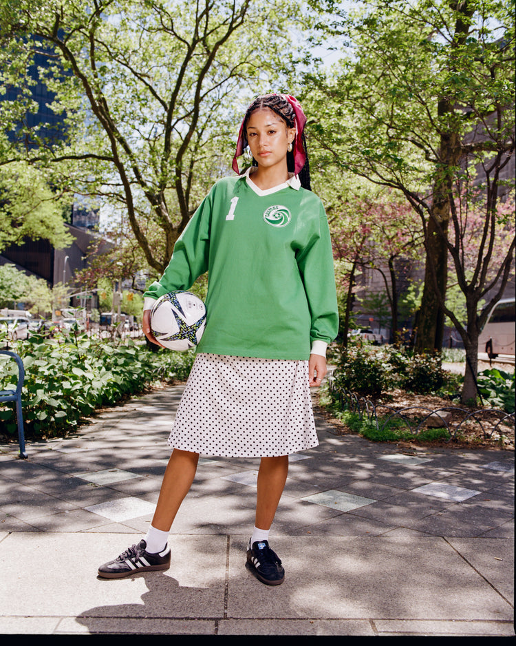 A young person stands outdoors in an Ebbets Field New York Cosmos Vintage Soccer Goalie Jersey, paired with a white polka-dot skirt, black sneakers, and white socks. They hold a soccer ball, have pink ribbons in braids, with trees and buildings behind them.
