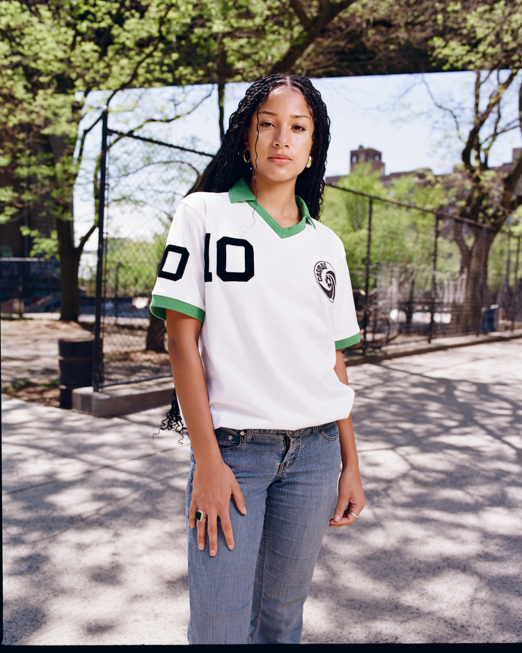 A woman with long braided hair stands outdoors on a sunny day, wearing the Ebbets Field New York Cosmos Vintage Collar Soccer Jersey with a green collar and number 10, paired with blue jeans. Trees and a metal fence are in the background.