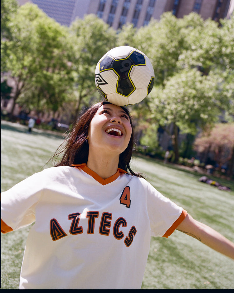 A smiling person wears an Ebbets Field Los Angeles Aztecs Vintage Collar Soccer Jersey and balances a soccer ball on their forehead in a sunny park with green trees and city buildings in the background.