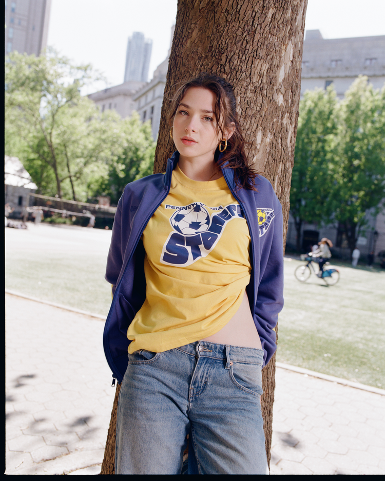A young woman in a park wears the Ebbets Field Pennsylvania Stoners Short Sleeve Graphic T-Shirt, paired with a blue jacket and jeans. Green trees, grass, urban buildings, and a passing cyclist suggest a sunny city day.