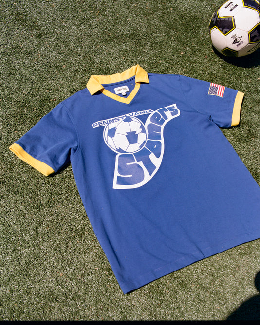 The Ebbets Field Pennsylvania Stoners Vintage Collar Soccer Jersey, in blue cotton with yellow collar and cuffs, features a vintage screen-printed logo, American flag patch on the sleeve, and is displayed lying on grass beside a soccer ball.