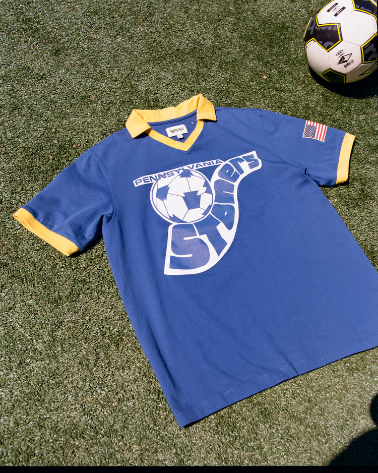The Ebbets Field Pennsylvania Stoners Vintage Collar Soccer Jersey, in blue cotton with yellow collar and cuffs, features a vintage screen-printed logo, American flag patch on the sleeve, and is displayed lying on grass beside a soccer ball.