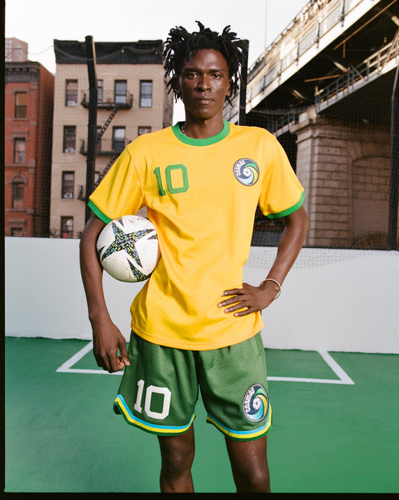 A soccer player stands on an outdoor court with a ball, wearing the yellow and green New York Cosmos Soccer Mesh Jersey by Ebbets Field and green shorts, with city buildings and an overpass in the background.