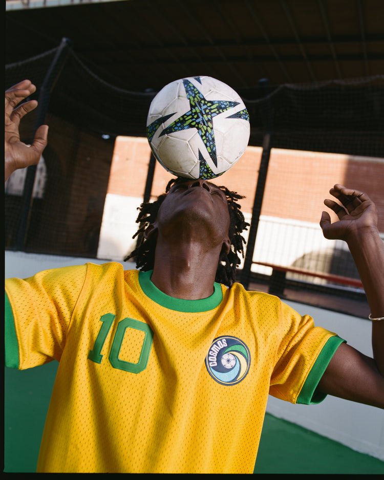 A soccer player in an Ebbets Field New York Cosmos Soccer Mesh Jersey with number 10 balances a ball on their forehead on an outdoor court, arms raised, with a brick wall and netting in the background.