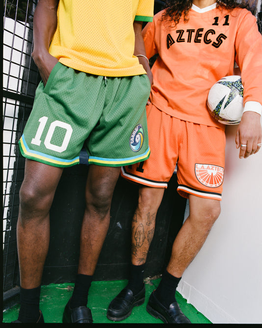Two people stand side by side in soccer apparel: one in green mesh shorts with “10” and a yellow jersey, the other holding a soccer ball, dressed in Ebbets Field Los Angeles Aztecs Mesh Soccer Shorts and an orange Aztecs jersey. Their heads are out of frame.