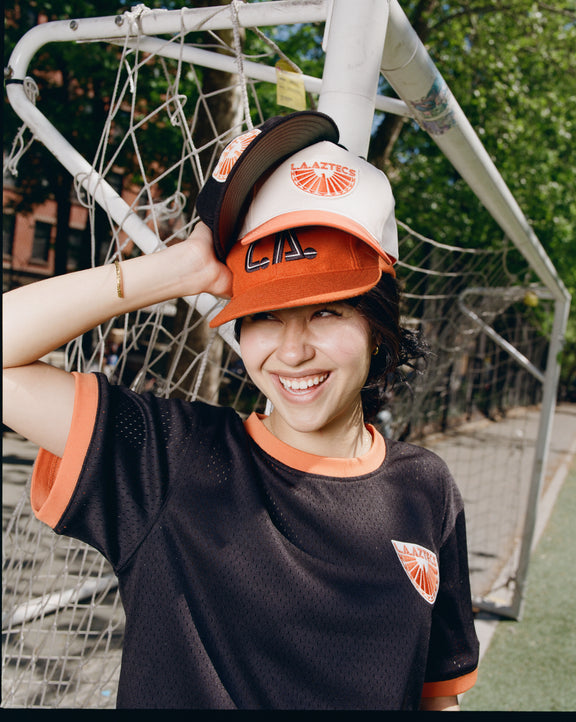 A smiling person in a black soccer jersey stands by the goalpost, stacking various hats— including the Ebbets Field Los Angeles Aztecs A-Frame Cotton Twill Ballcap— on their head. Trees and a soccer field are visible in the background.