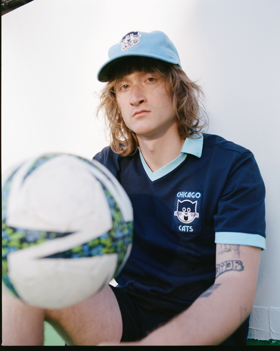 Wearing the Ebbets Field Chicago Cats Vintage Collar Soccer Jersey, a long-haired individual pairs it with a matching cap, sitting before a white wall with a soccer ball—an ideal look for American Soccer League fans.