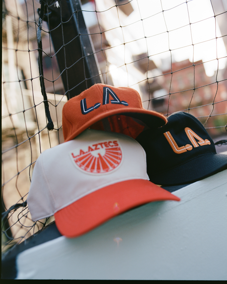 Three Ebbets Field Los Angeles Aztecs Vintage Wool Ballcaps sit on a ledge by a net fence—two orange with “L.A.” logos, one white and orange with the Aztecs logo, nodding to the NASL era. Urban buildings blur in the background.