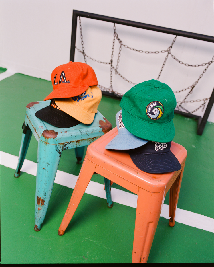 Two colorful metal stools hold six stacked baseball caps, including the Ebbets Field New York Cosmos Wool Ballcap, arranged on a green indoor court with a soccer net in the background—a tribute to Pelé’s legacy.
