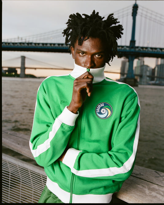 Someone stands by a waterfront railing at dusk, partially covering their face with the collar of an Ebbets Field New York Cosmos Soccer Warm Up Jacket. Bridges and a city skyline are visible in the background.