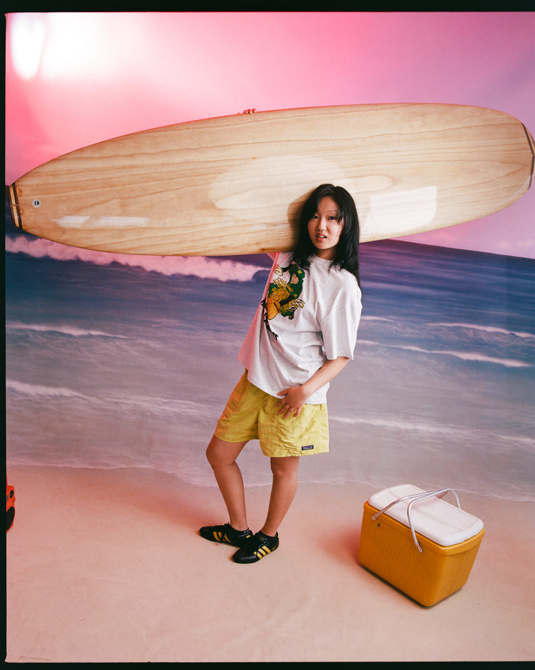 Indoors, a person stands before a beach-themed backdrop with a wooden surfboard on their shoulder, wearing an Ebbets Field Hawaii Islanders (PCL) Short Sleeve Graphic T-Shirt, yellow shorts, black shoes, next to a yellow cooler.