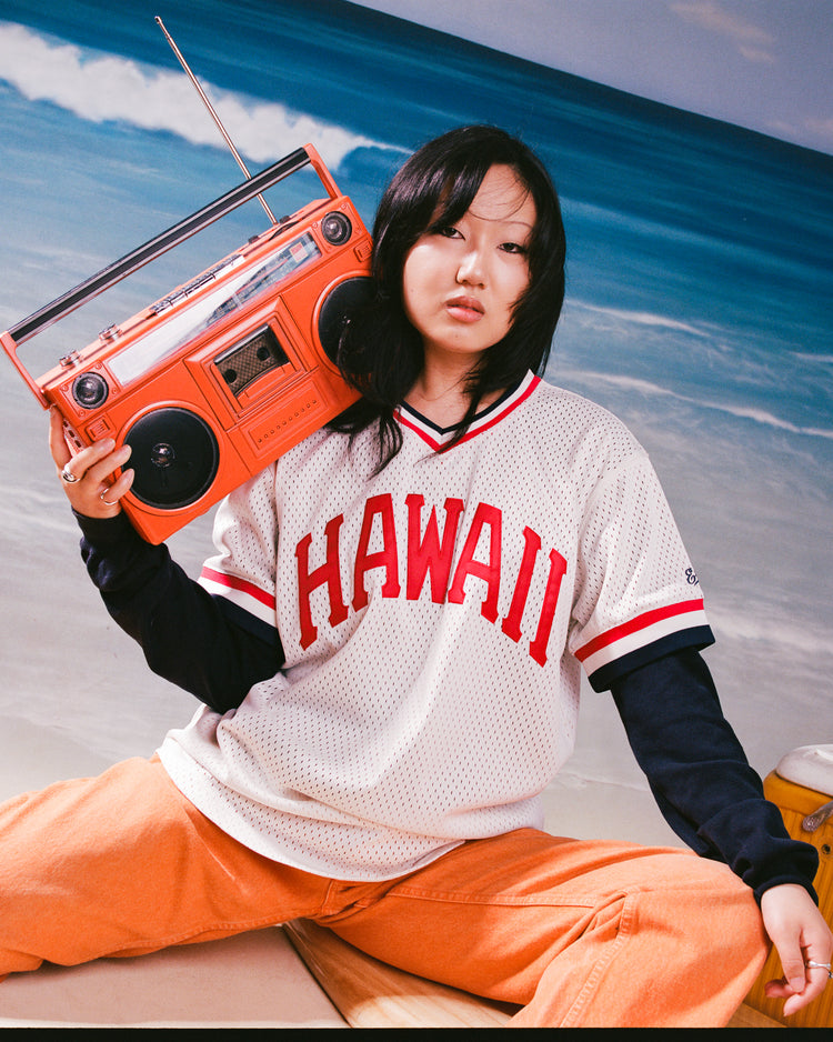 Wearing Ebbets Field's Hawaii Islanders (PCL) 1974 Mesh V-Neck Jersey and orange pants, a person sits on the sand with a red boombox by their ear, a yellow cooler beside them, and the ocean in the background.