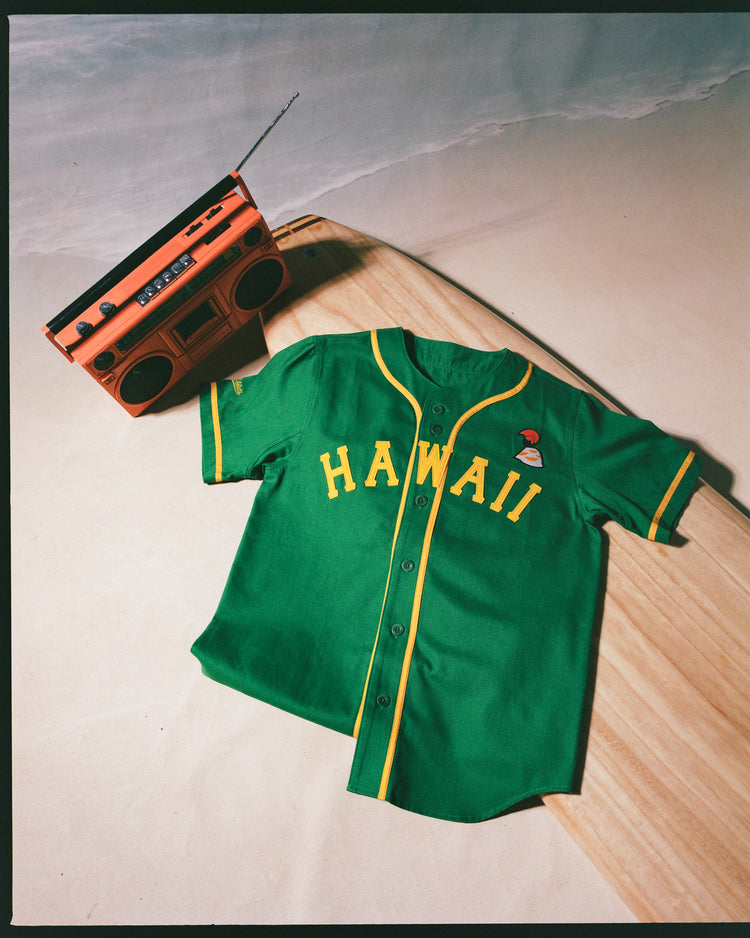 The Ebbets Field Hawaii Islanders (PCL) Cotton Twill Baseball Jersey in green with yellow trim and HAWAII on the front rests on a wooden surfboard beside an orange vintage boombox, set against a sand-colored backdrop for classic Pacific Coast League vibes.