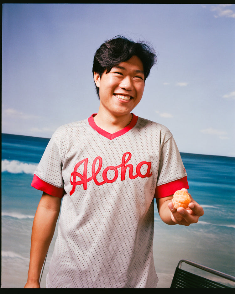 A smiling person wears the Ebbets Field Aloha 100th Infantry 1942 Mesh V-Neck Jersey, gray and red, while standing before a beach backdrop and holding a peeled orange segment.