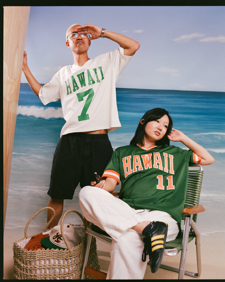 Two people pose by a beach backdrop. One stands holding a board in a white Ebbets Field University of Hawaii Rainbow Warriors Mesh Football Jersey and black shorts. The other sits in a green Rainbow Warriors jersey, white pants, and black sneakers beside a basket.