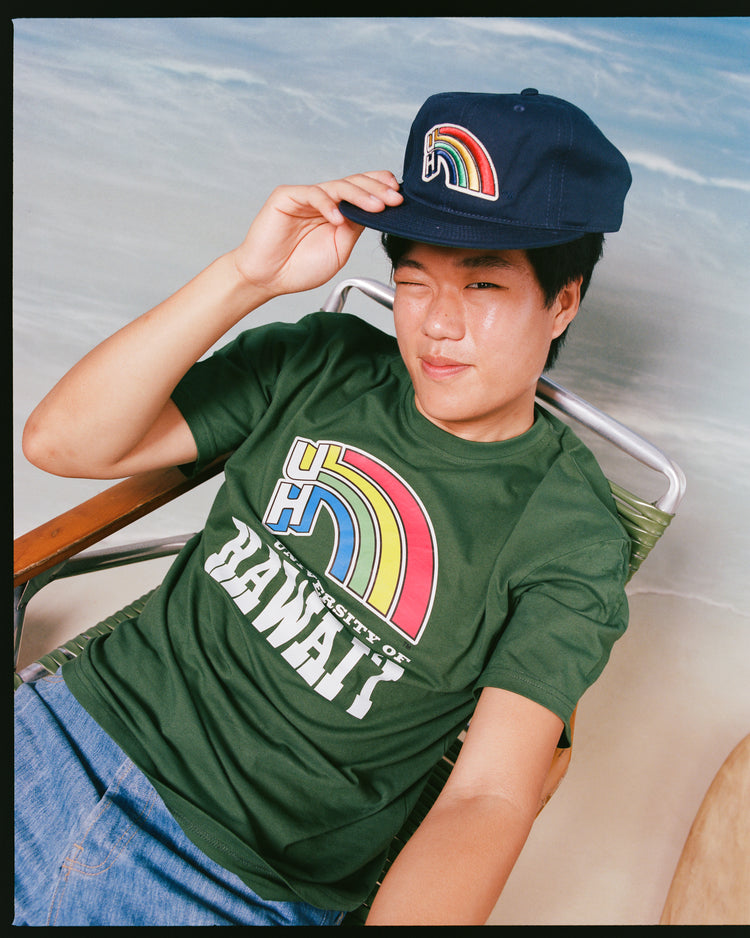 A person sits on a chair in blue jeans, wearing an Ebbets Field University of Hawaii Rainbow Warriors Short Sleeve Graphic T-Shirt and holding a navy cap with the rainbow logo, set against a beach-themed background.