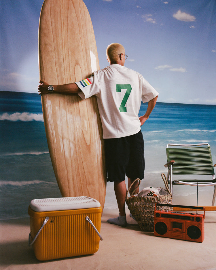 A blond person stands indoors before a beach backdrop holding a surfboard, wearing Ebbets Field’s University of Hawaii Rainbow Warriors Mesh Football Jersey, dark shorts, and sunglasses. A yellow cooler, retro radio, and beach chair are nearby.