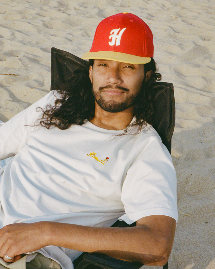 A man with long curly hair and a beard relaxes on a beach chair, wearing the Ebbets Field Hawaii Islanders (PCL) Graphic Short Sleeve Tee and a red cap with a white H.