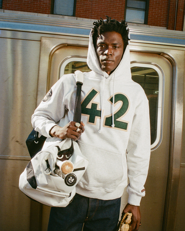 Wearing an Ebbets Field Karabo Poppy x Ebbets Field Jackie Robinson Heavyweight Cotton Pullover Hoodie, a person stands by a subway train, holding a white bag with pins and patches and a small brown object in the other hand.