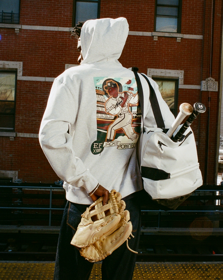 Facing away on a train platform, a person wears the Ebbets Field Karabo Poppy x Jackie Robinson Heavyweight Cotton Pullover Hoodie, carries a white backpack with bats, and holds a tan glove. A brick building behind honors Black History Month.