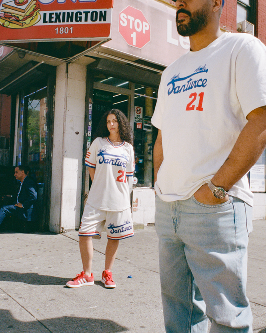 Two people wearing matching white Santurce 21 shirts stand on a city sidewalk near a storefront at Lexington and 1801. A man sits in the background, and a stop sign is visible above the scene.