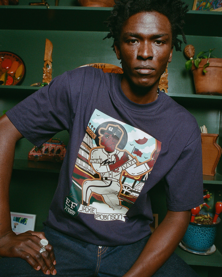 A man sits indoors wearing the Karabo Poppy x Ebbets Field Jackie Robinson Short Sleeve Graphic Tee by Ebbets Field, featuring a colorful illustrated player with a baseball field background. Shelves with plants and decor are behind him.