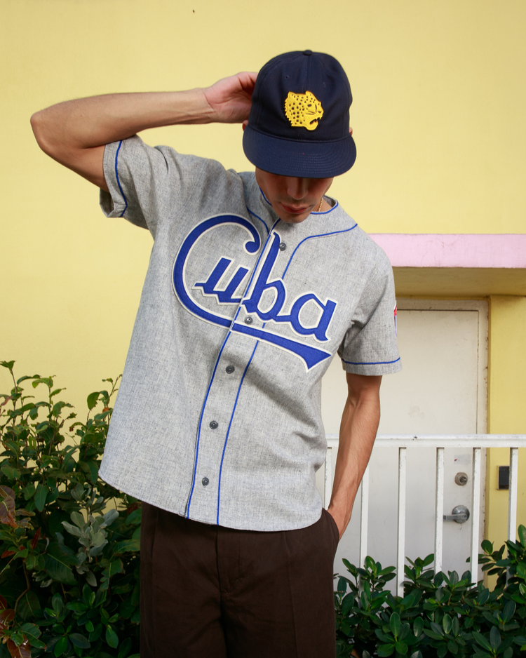 A man in an EFF Cuba National Team 1947 Authentic Wool Road Baseball Jersey, gray with blue lettering, and a navy cap with a yellow patch stands before a pale yellow wall and white railing, looking down as he adjusts his hat.
