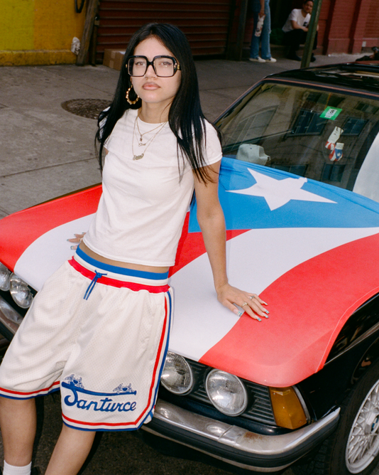 A woman with long dark hair, glasses, and layered necklaces leans against a car with a Puerto Rican flag design on its hood. She wears a white T-shirt and basketball shorts that say Santurce. The scene is urban.