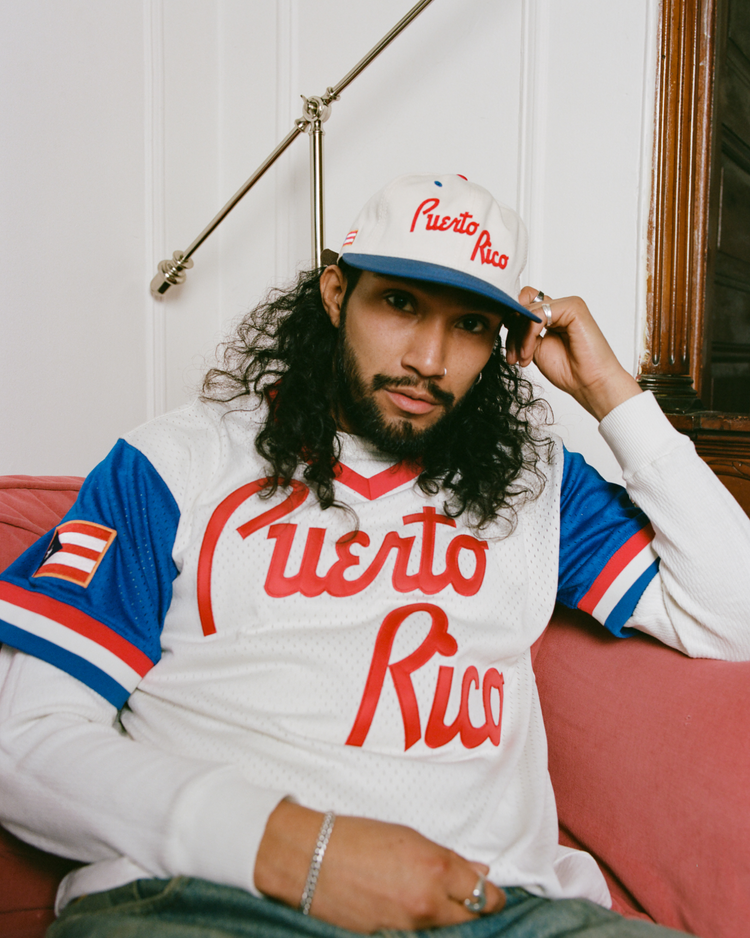 A person with long curly hair and a beard sits thoughtfully on a pink couch, wearing a white and blue Puerto Rico 1958 National Team Cotton Twill Ballcap by Ebbets Field, evoking Roberto Clemente's quiet intensity.