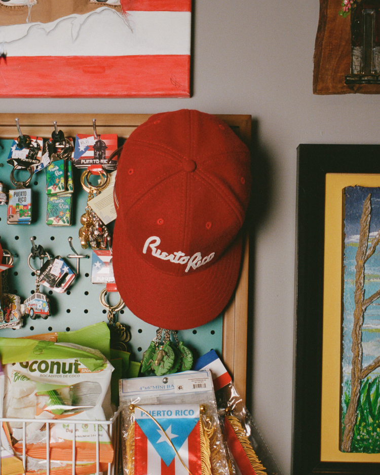 An Ebbets Field Puerto Rico 1958 National Team Wool Ballcap hangs on a pegboard, surrounded by keychains, a Puerto Rican flag, snacks, and colorful framed artwork.